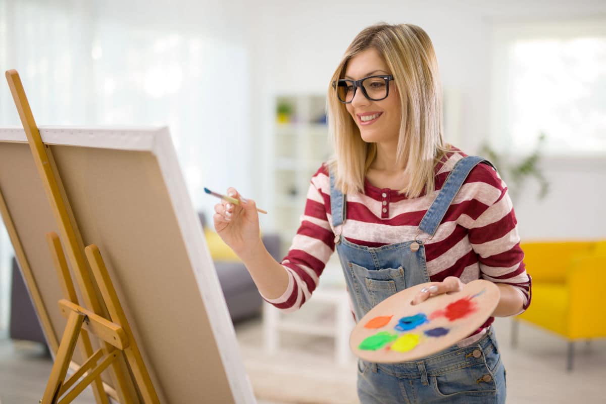 Young female artist painting on a canvas with a paintbrush and holding a color palette at home